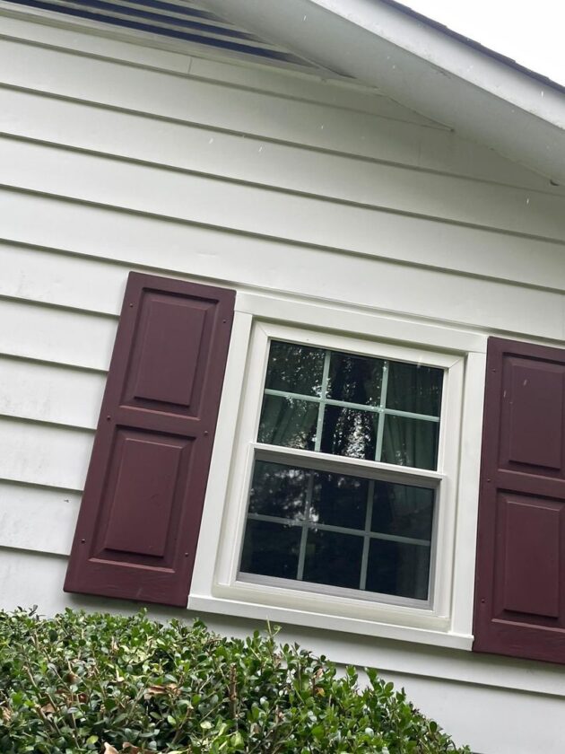 Close-up of a new double-hung window with white frame and dark red shutters on a house with white siding