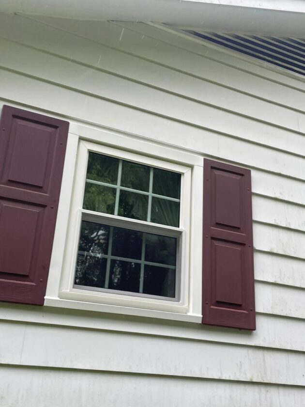 Close-up of a new double-hung window with maroon shutters on a house's white siding