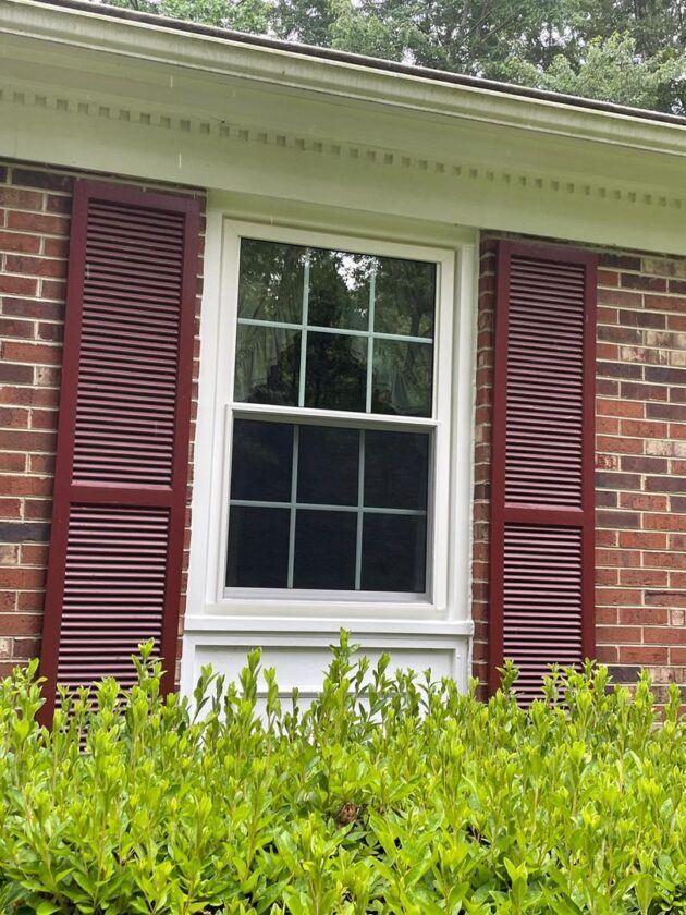 A new double-hung window with white trim and red shutters on a brick house, surrounded by green shrubs