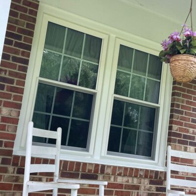 Two new white double-hung windows on a brick house porch with white rocking chairs and a hanging flower basket