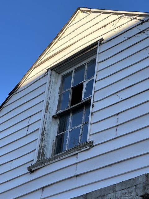 Upward view of a damaged window with peeling paint on a house's white siding against a blue sky