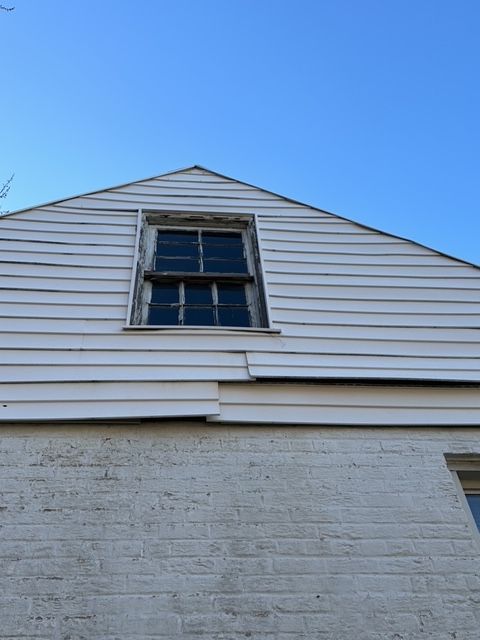 Upper level window with peeling paint on a house with white siding and a clear blue sky