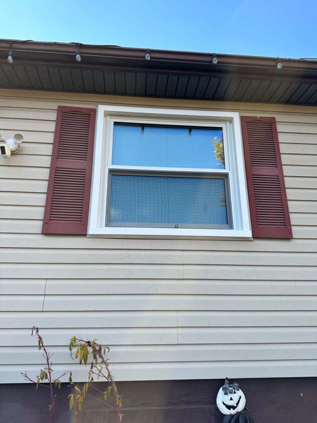 Exterior view of a house with a new double-hung window with white frames and maroon shutters against beige siding