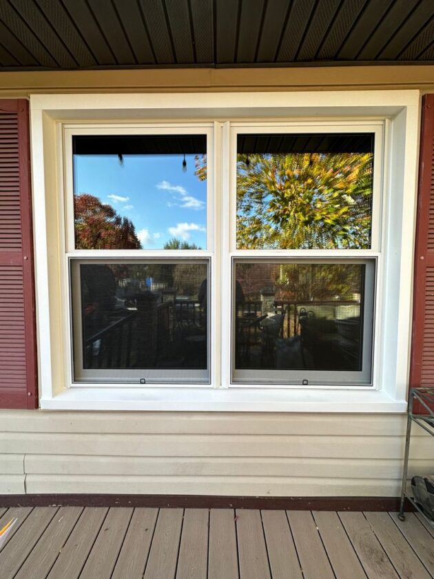 Two new white double-hung windows on the side of a house with reflections of trees and sky