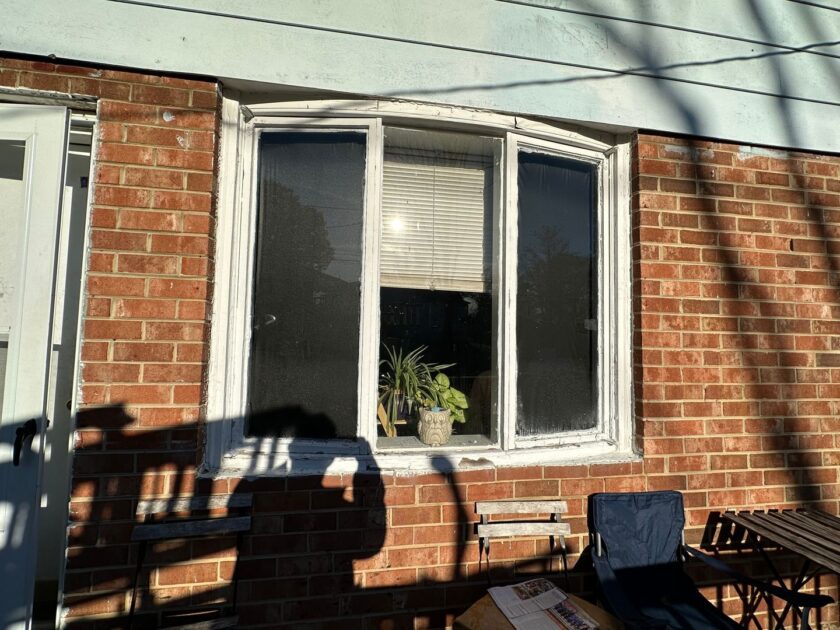 Old white-framed window on a brick house with a potted plant on the sill and shadows of a person and patio furniture