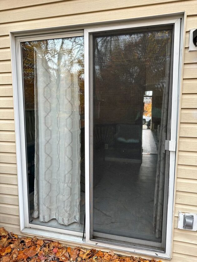 Sliding patio door with a white curtain and screen, set in a beige siding wall with fallen leaves on the ground